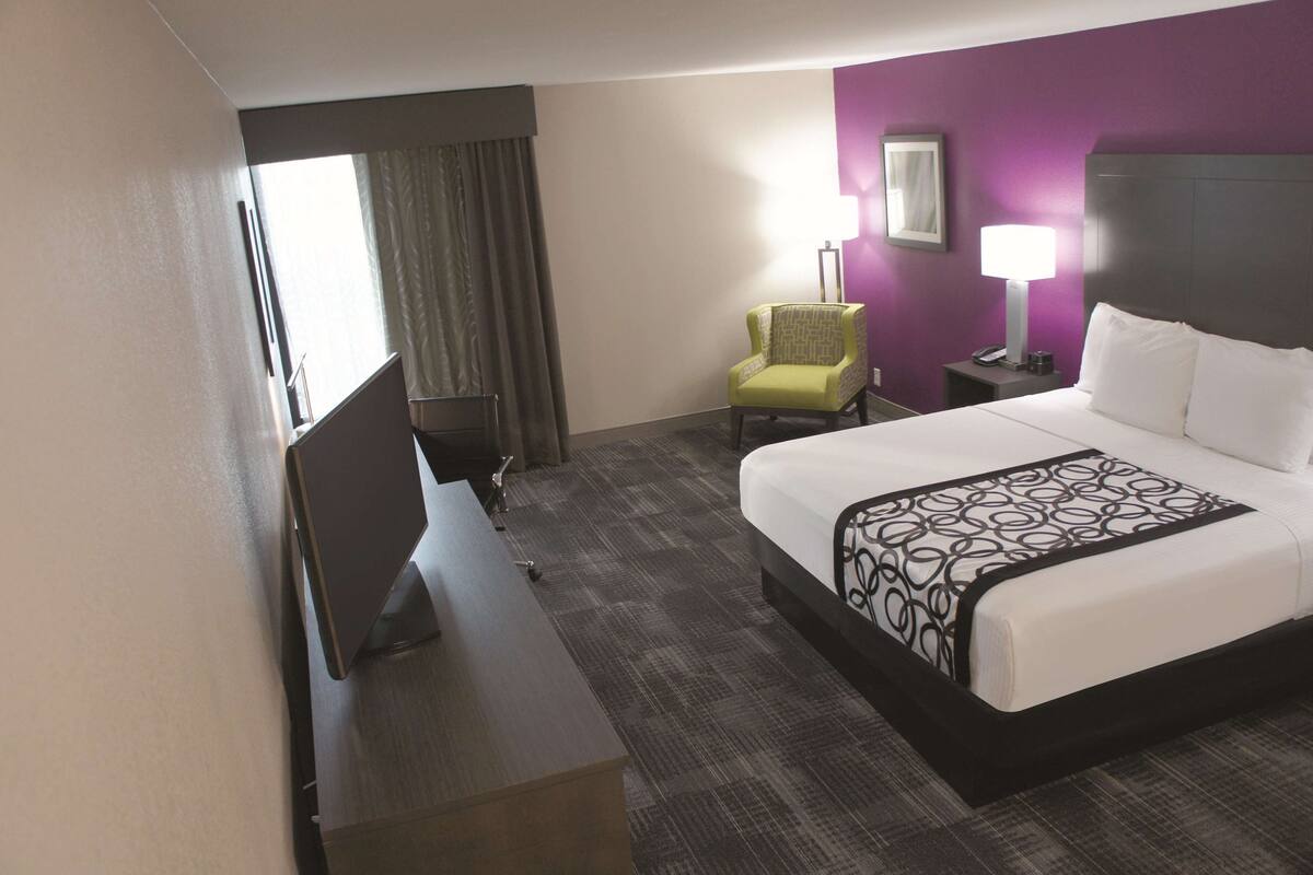 A hotel room with a bed, a desk, a chair, a TV, and a nightstand with a lamp.