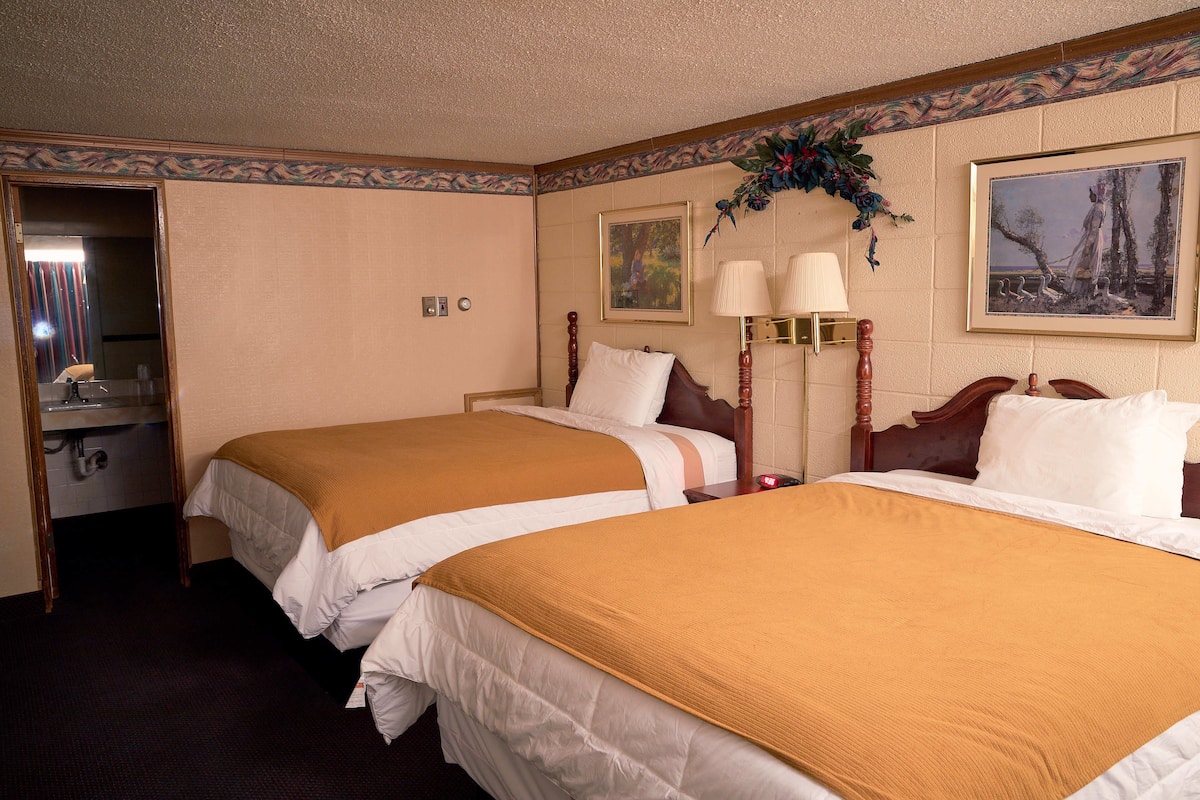 A hotel room with two beds, a wooden headboard, a mirror, a desk with a lamp, and a framed painting on the wall.