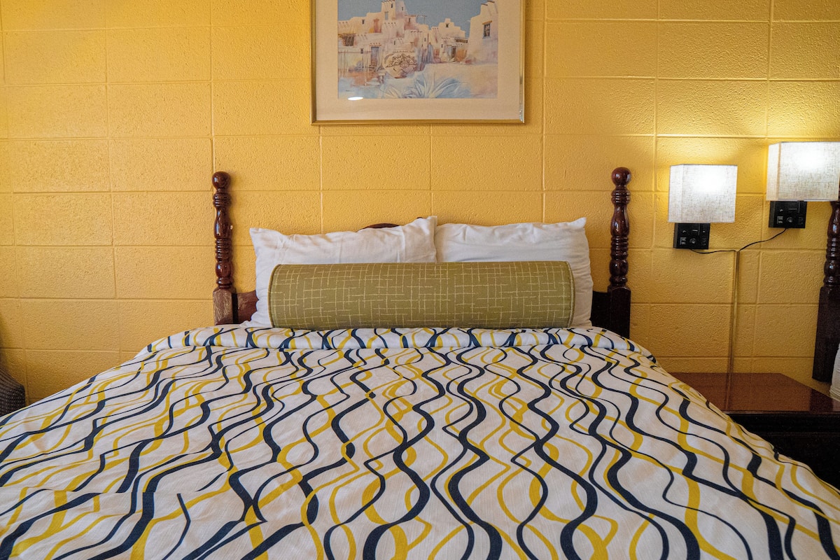 A hotel room with a bed, a framed picture on the wall, and two wall-mounted lamps.