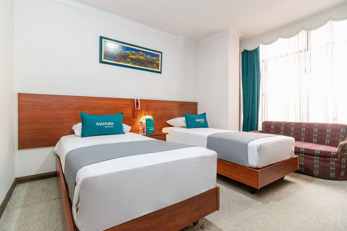 A hotel room with two beds, a wooden headboard, a nightstand with a lamp, a red plaid sofa, and a window with curtains.