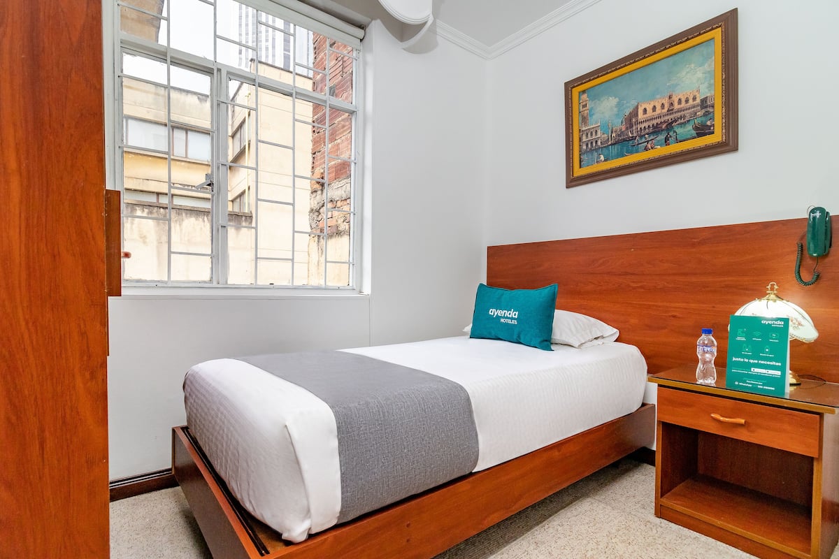 A hotel room with a bed, a nightstand, a framed picture, and a window with a view of buildings.