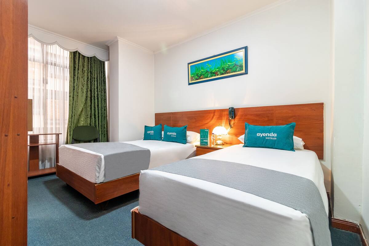 A hotel room with two beds, each with a wooden headboard, white bedding, and teal pillows labeled 'ayenda hotels'. There is a window with sheer curtains, a framed picture on the wall, and a small table with a lamp.