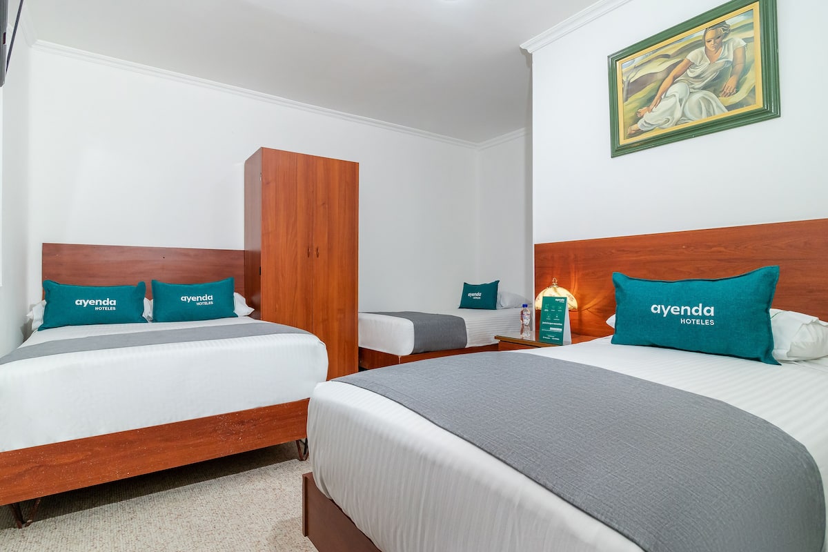 A hotel room with two beds, each with a wooden headboard, white bedding, and teal pillows with 'ayenda hotels' branding.