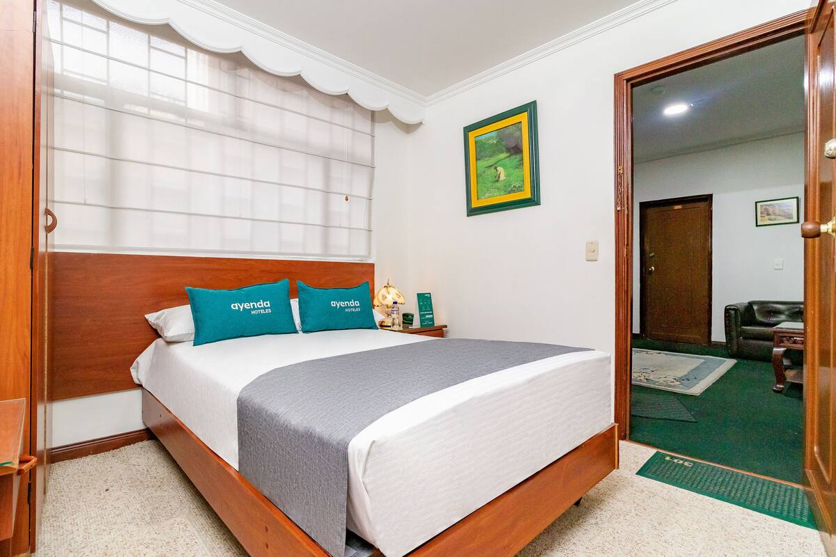A hotel room with a wooden bed, white bedding, teal pillows, a green and yellow framed picture, and a wooden door.