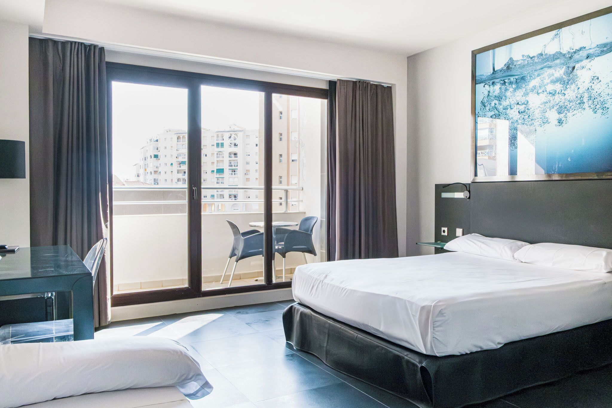 Habitación de hotel con una cama grande, un escritorio, una silla y vistas a edificios a través de una puerta corredera.