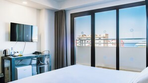 Habitación de hotel con una cama, un escritorio con computadora, televisor y vistas a la ciudad a través de amplios ventanales.