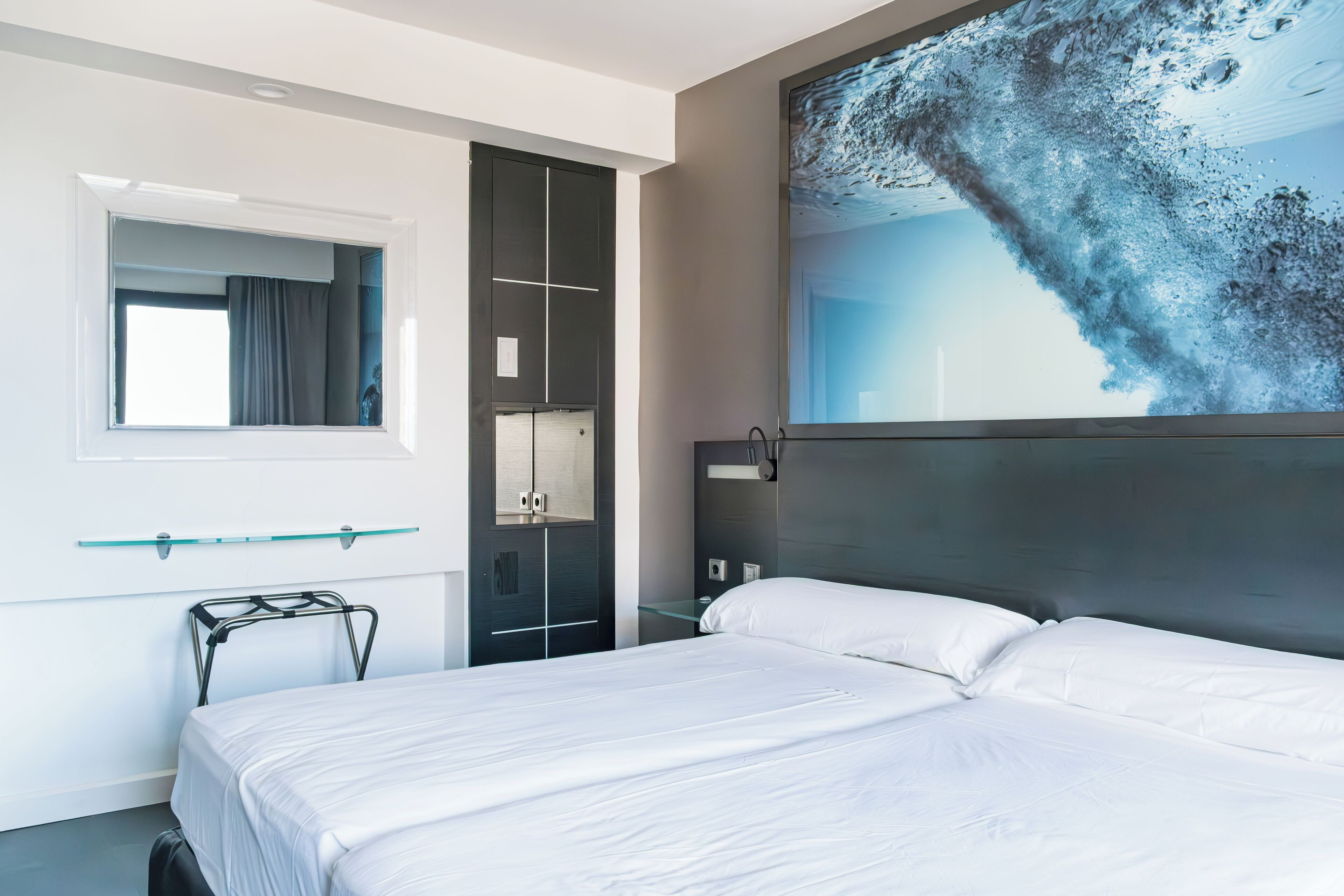 Una habitación de hotel moderna con una cama grande, un estante de vidrio, un armario y un cuadro abstracto de gran tamaño en la pared.