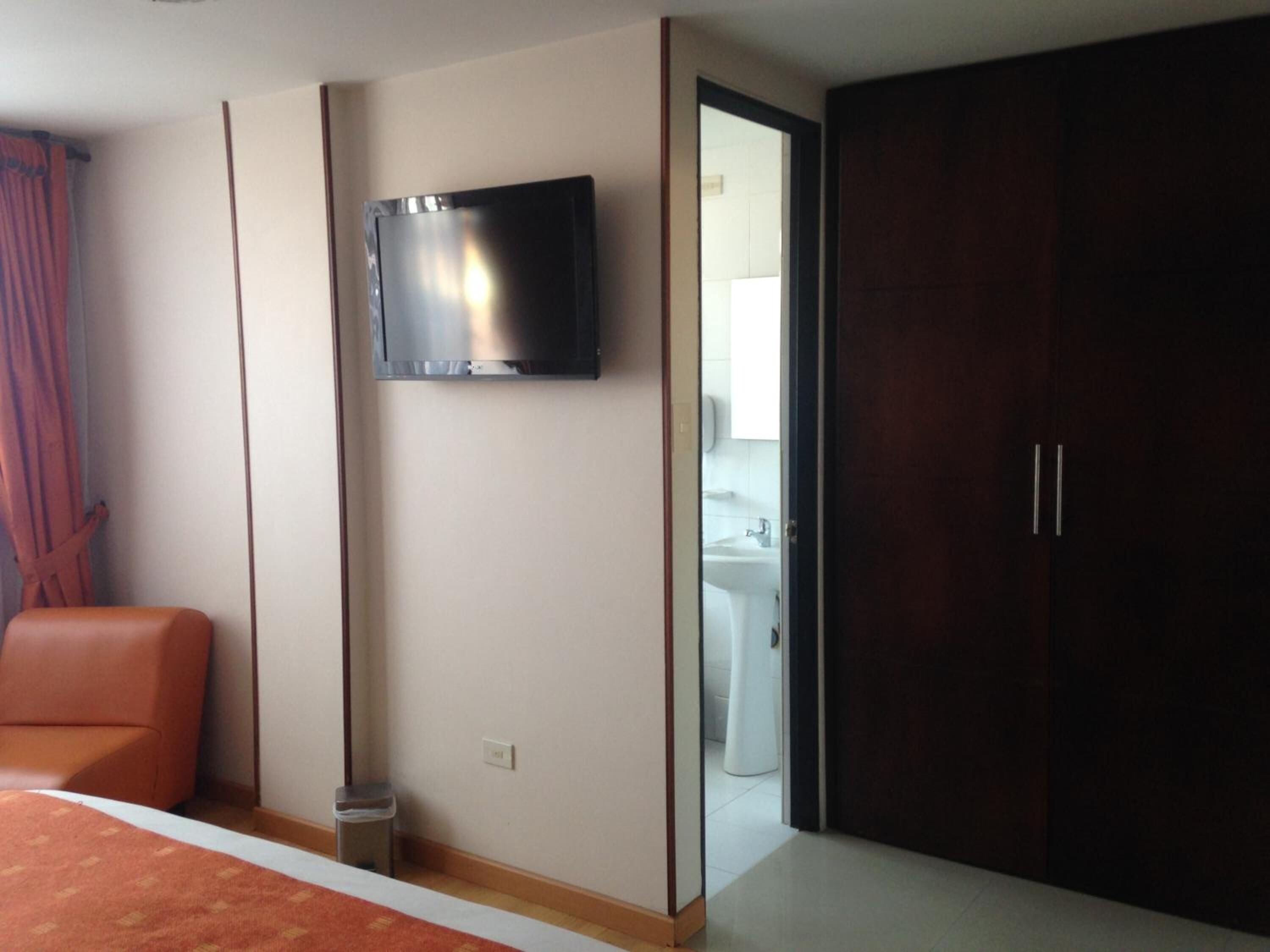 Habitación de hotel con cama, televisor montado en la pared, armario y baño visible a través de una puerta abierta.