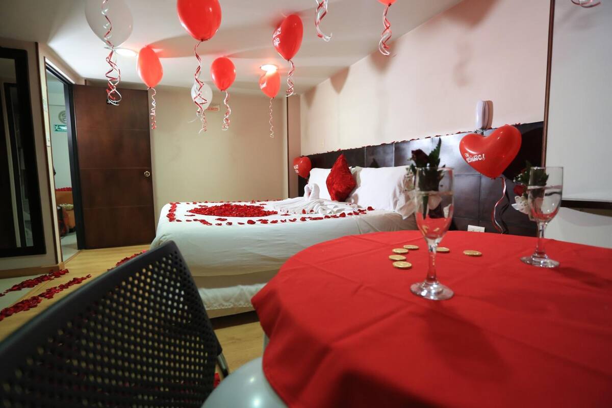 Ein Hotelzimmer mit einem Bett, das mit roten Rosenblättern dekoriert ist, einem roten Tisch mit Weingläsern und herzförmigen Ballons.