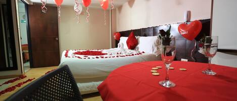 Habitación de hotel con una cama decorada con pétalos de rosa roja, una mesa roja con copas de vino y globos con forma de corazón.