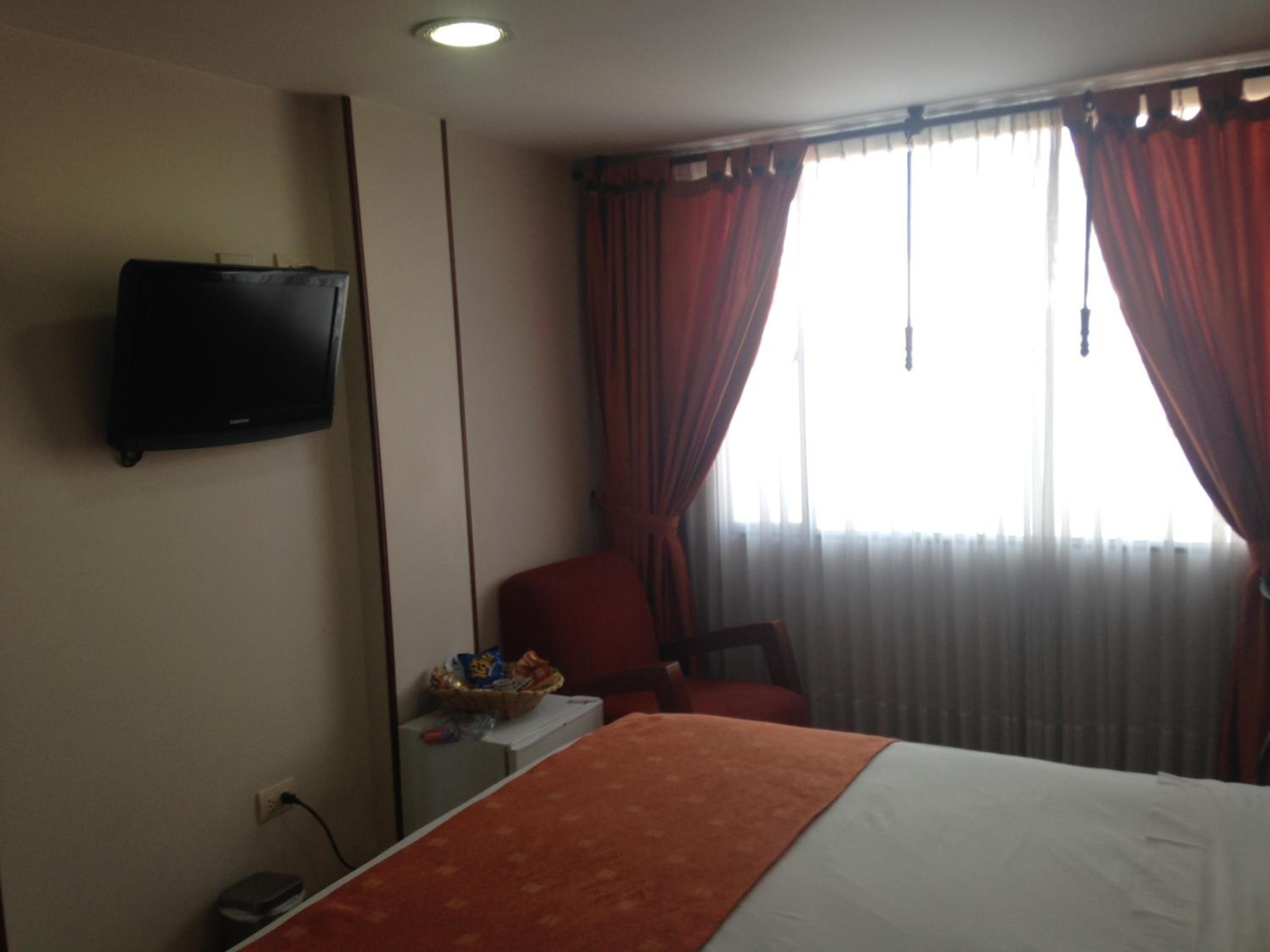 A hotel room with a bed, a television mounted on the wall, a chair, a small table, and a window with curtains.