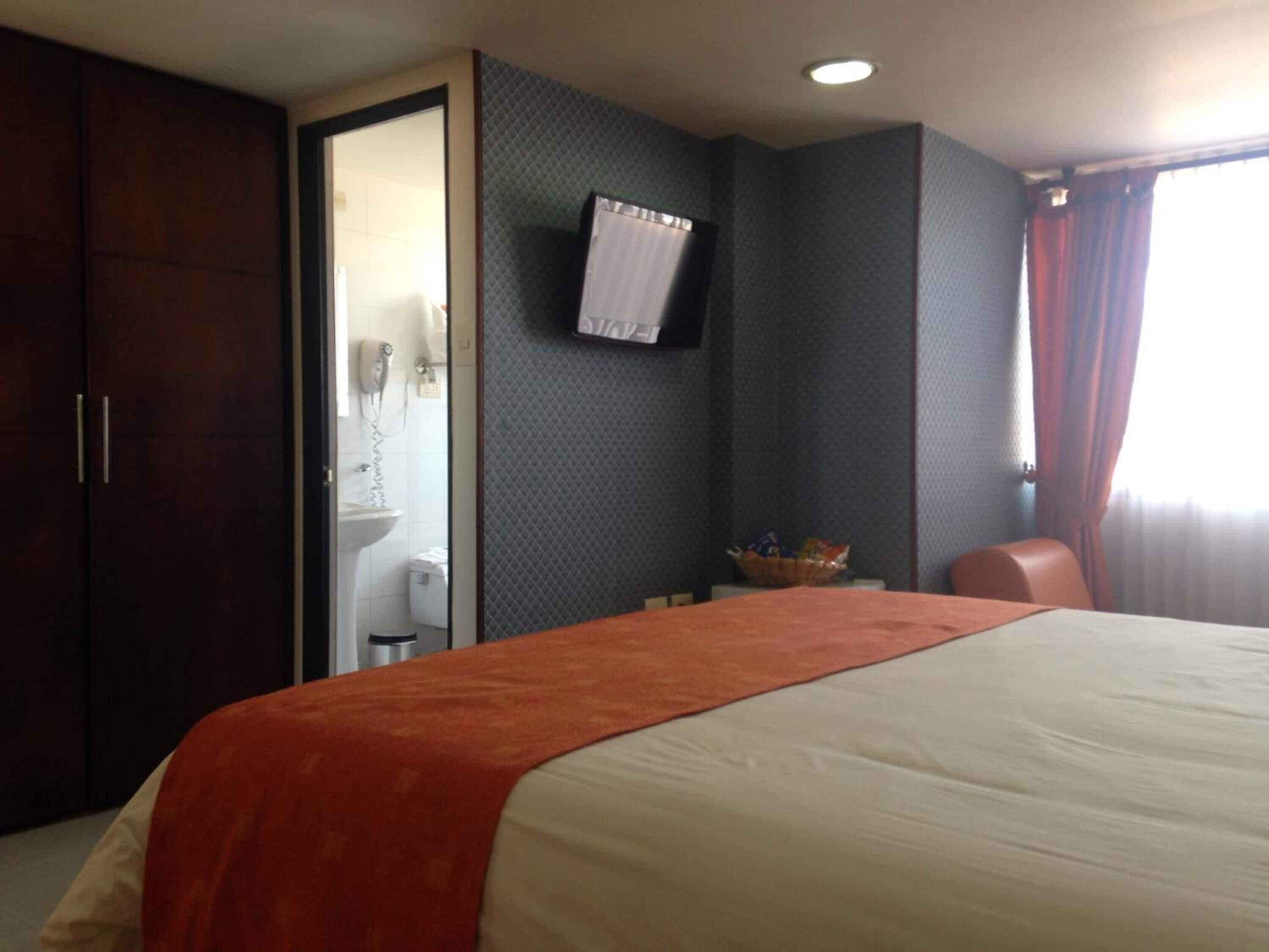 A hotel room with a bed, a television mounted on the wall, a bathroom visible through an open door, and a window with curtains.