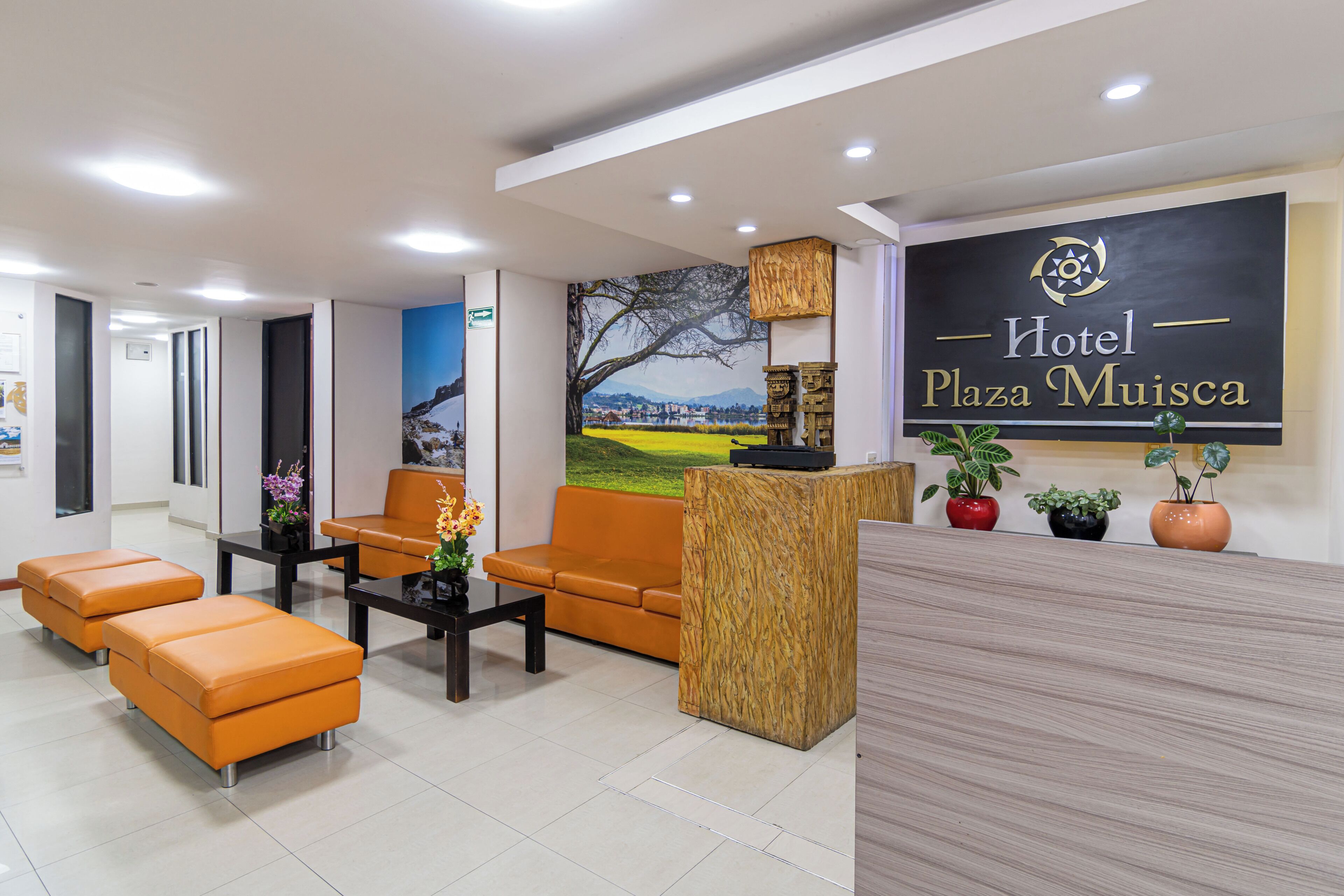 A hotel lobby with a reception desk, orange seating, a large wall mural, and a sign reading 'Hotel Plaza Muisca'.