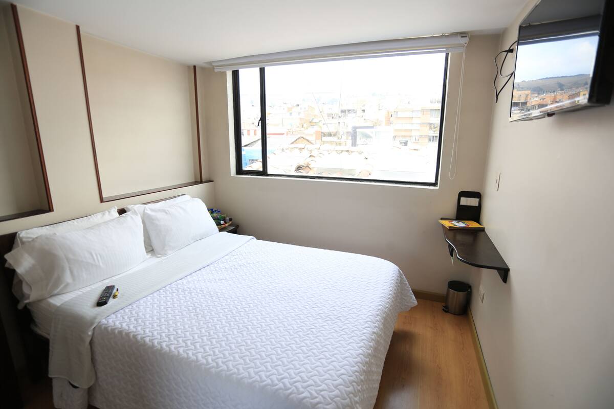 Ein Hotelzimmer mit einem großen Bett, einem Fenster mit Blick auf die Stadt, einer kleinen Ablagefläche und einem Mülleimer.
