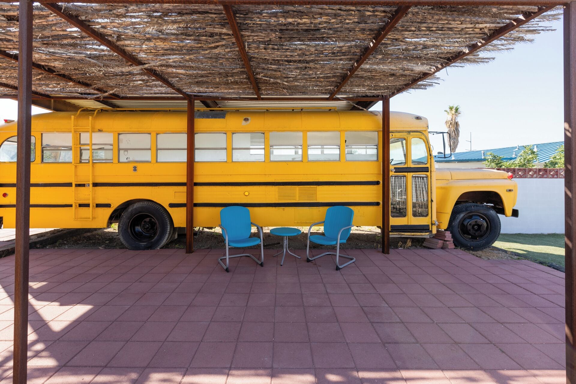 A yellow school bus parked under a thatched roof structure with two blue chairs in front.