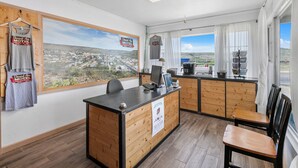 A small, well-lit room with a wooden desk, a computer, and a view of a town through large windows. - Desert Air Motel (Sanderson)