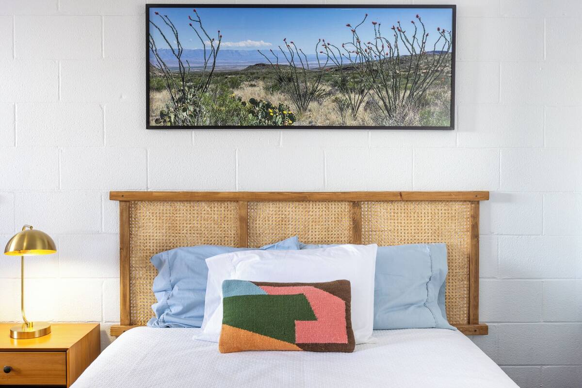 A bed with a wooden headboard, a nightstand with a lamp, and a framed picture of a desert landscape on the wall.