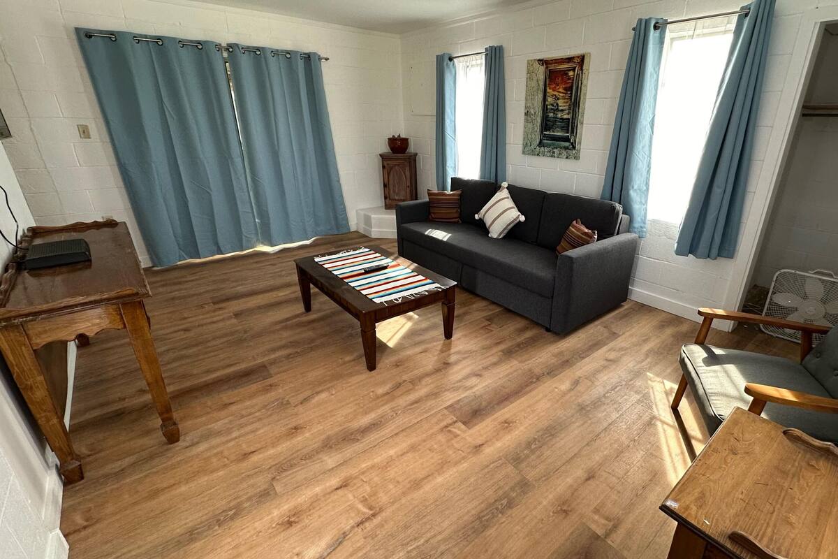 A living room with a sofa, coffee table, and a chair. There is a wooden floor and a window with curtains.
