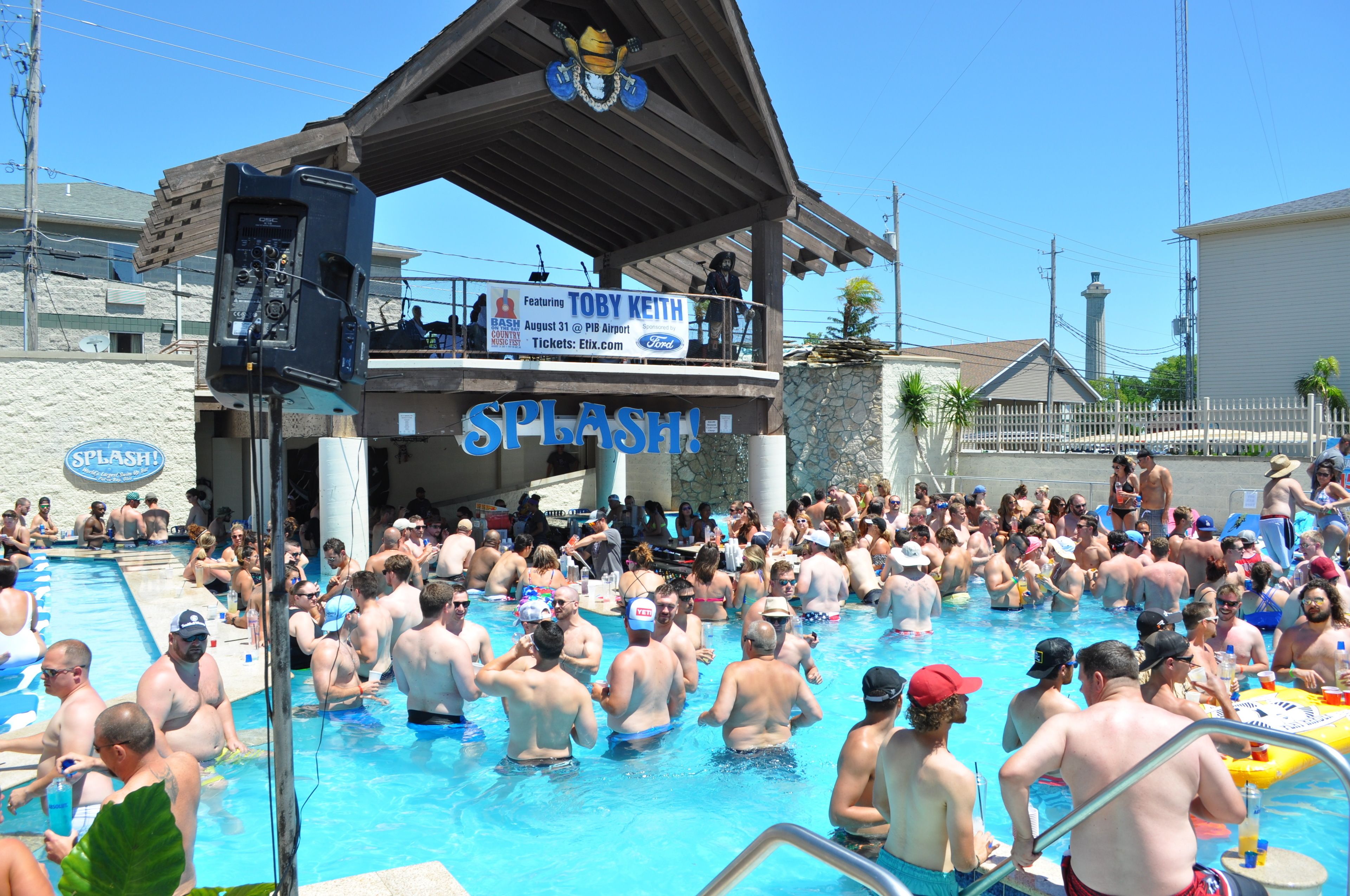 A crowded outdoor swimming pool with a stage and a sign that reads "SPLASH!".