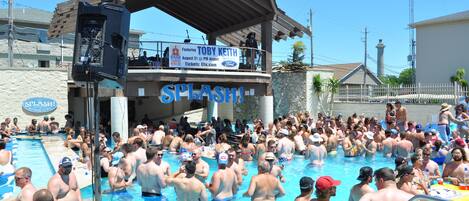 A crowded outdoor swimming pool with a stage and a sign that reads "SPLASH!".
