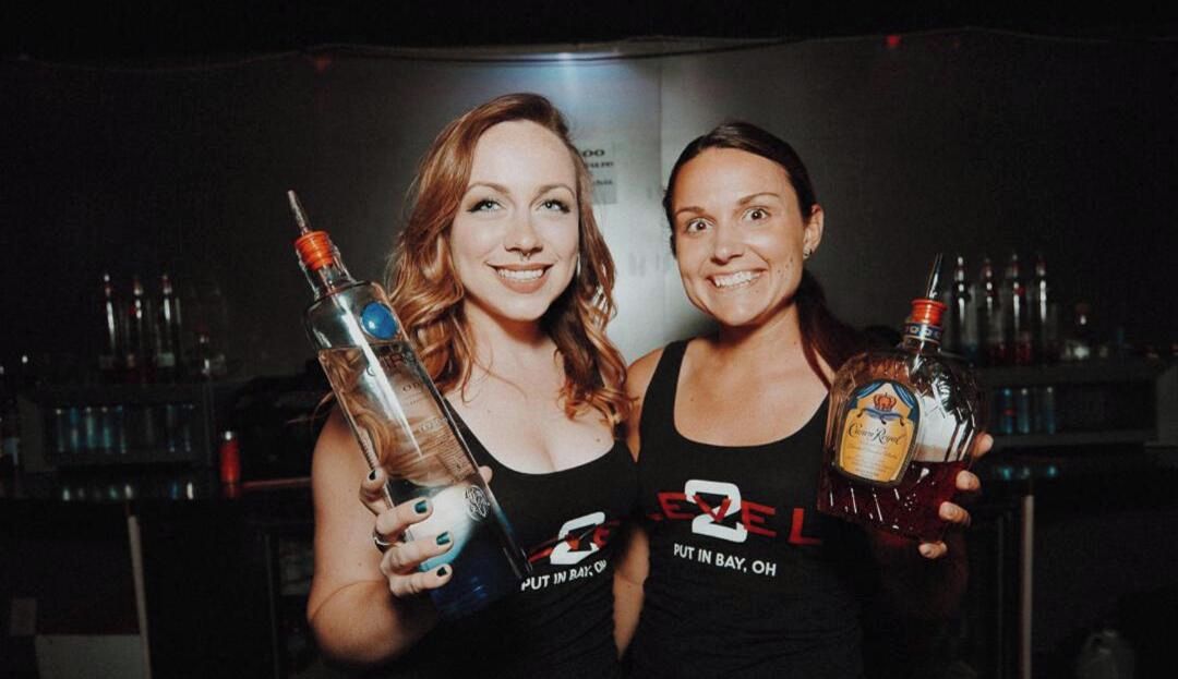 Dos personas sostienen botellas de licor en un ambiente con poca luz, con un bar al fondo.