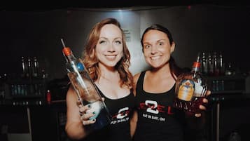 Two individuals holding bottles of liquor in a dimly lit setting with a bar in the background.