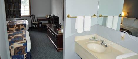 A hotel room with a bed, a desk, a dresser, a large mirror, and a bathroom with a sink and towel rack.