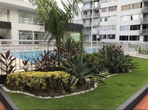 A modern hotel with a swimming pool surrounded by well-maintained greenery and a white fence.