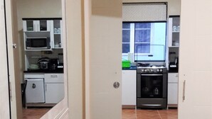 A compact kitchen with a microwave, stove, and cabinets. A bed with a checkered blanket is visible in the foreground.
