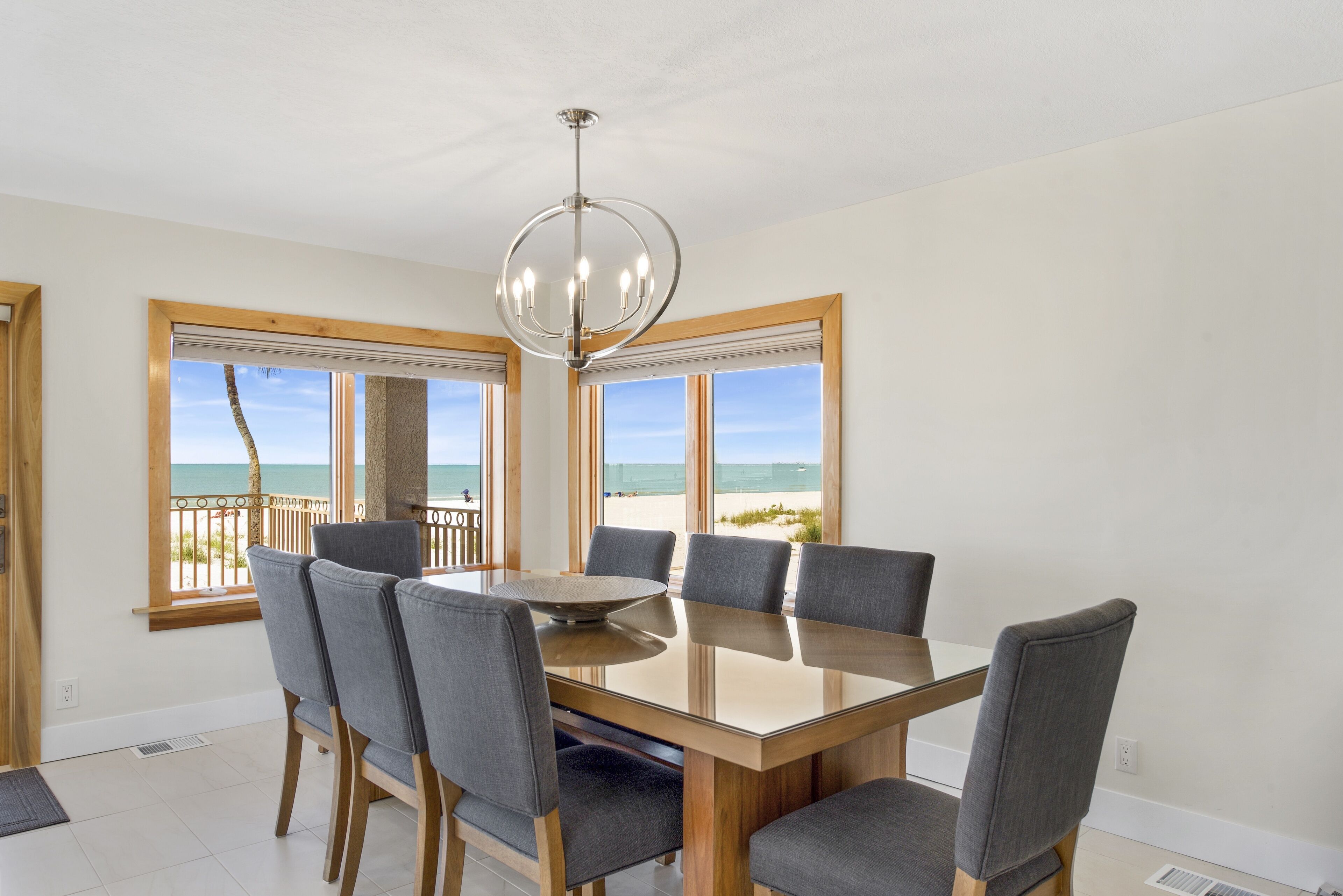 A dining area with a glass table and six chairs, large windows with a view of the beach, and a modern chandelier.