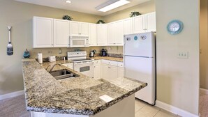 Una cocina con gabinetes blancos, encimeras de granito, microondas, estufa y refrigerador.