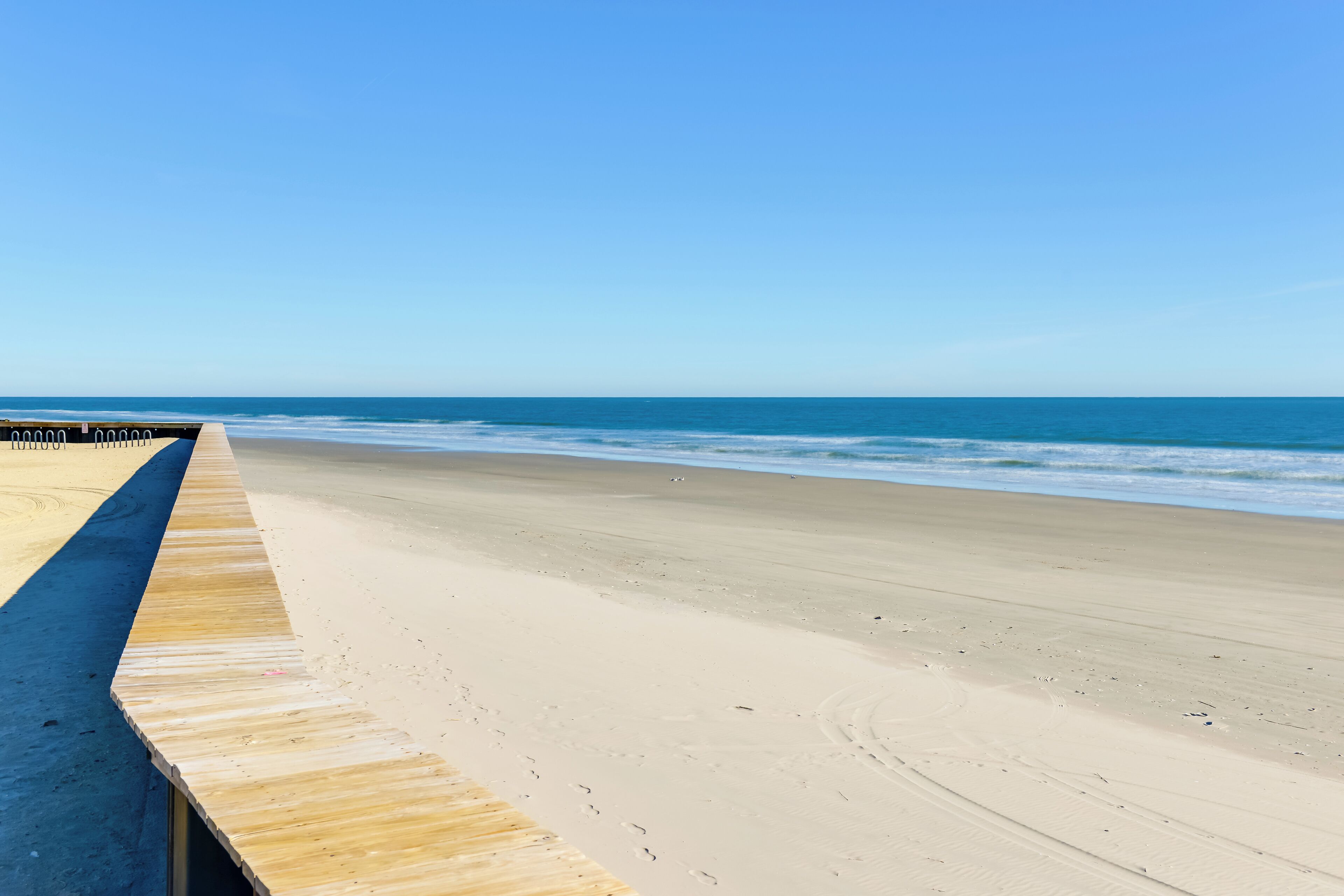Un paseo marítimo de madera que conduce a una playa arenosa, con huellas y un cielo azul despejado.
