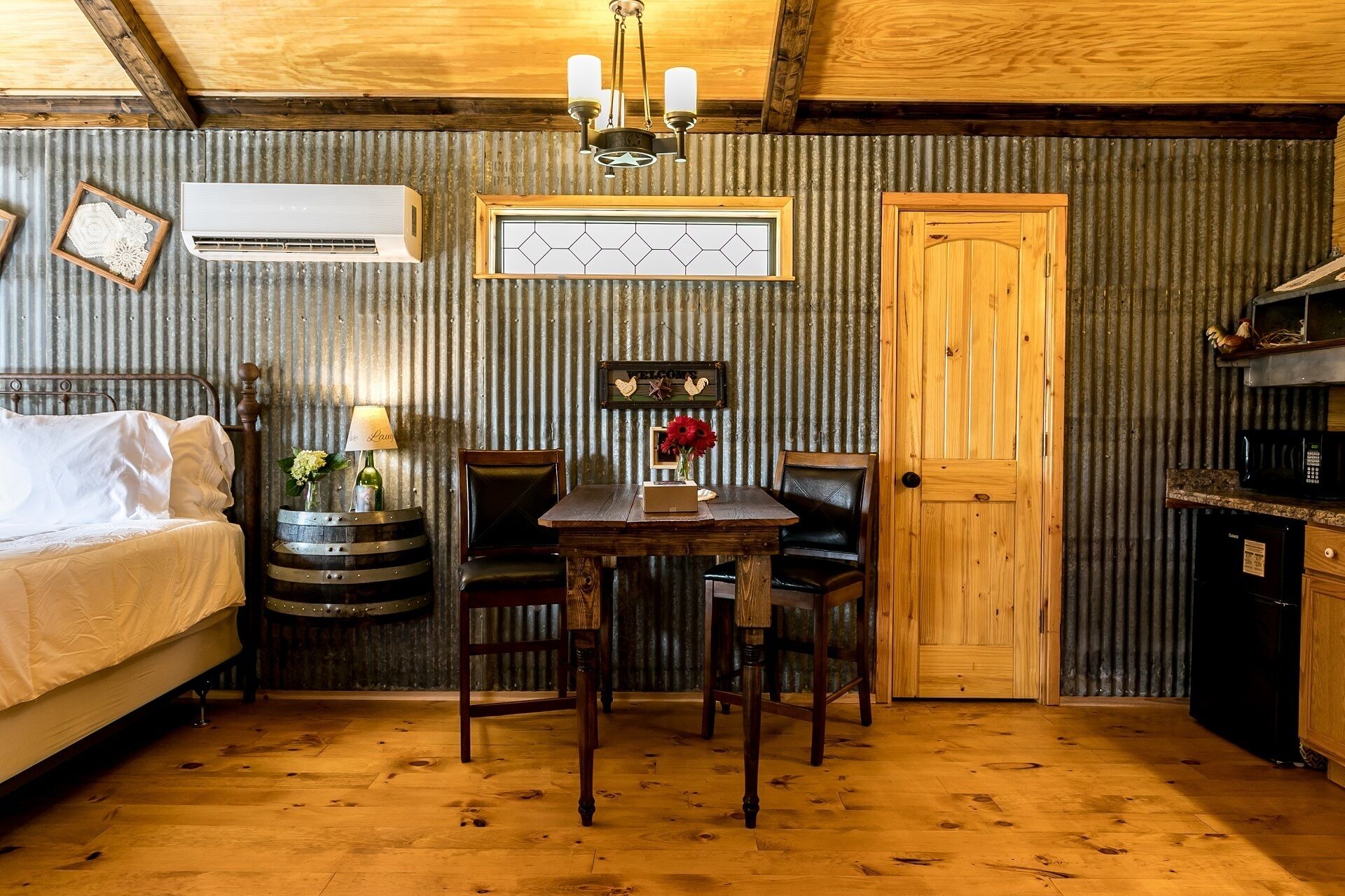 Una habitación rústica con una cama de madera, una mesa de comedor con sillas, una pequeña cocina y una puerta de madera.