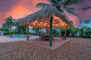 A thatched gazebo with a pool and patio furniture in a landscaped outdoor area. - Grande Palermo – Come stay and experience coastal living at its best! (Fort Myers Beach)