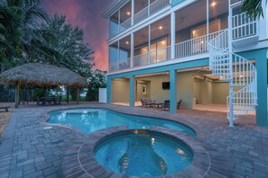 A two-story building with a swimming pool, outdoor furniture, and a thatched umbrella.