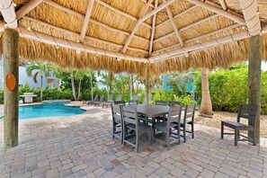 An outdoor pool area with a thatched roof, lounge chairs, and a dining table.