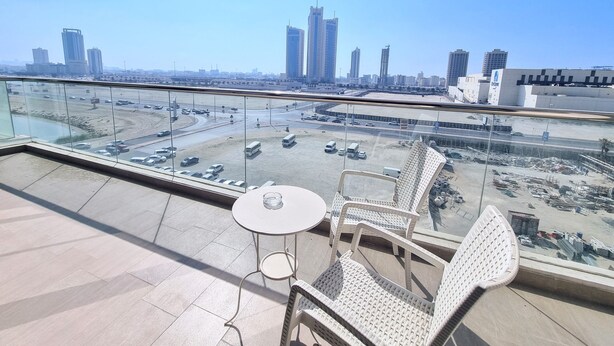 Una terraza en la azotea con barandilla de cristal, una mesa redonda y sillas de mimbre. Se ve un paisaje urbano con rascacielos y una obra en construcción al fondo.