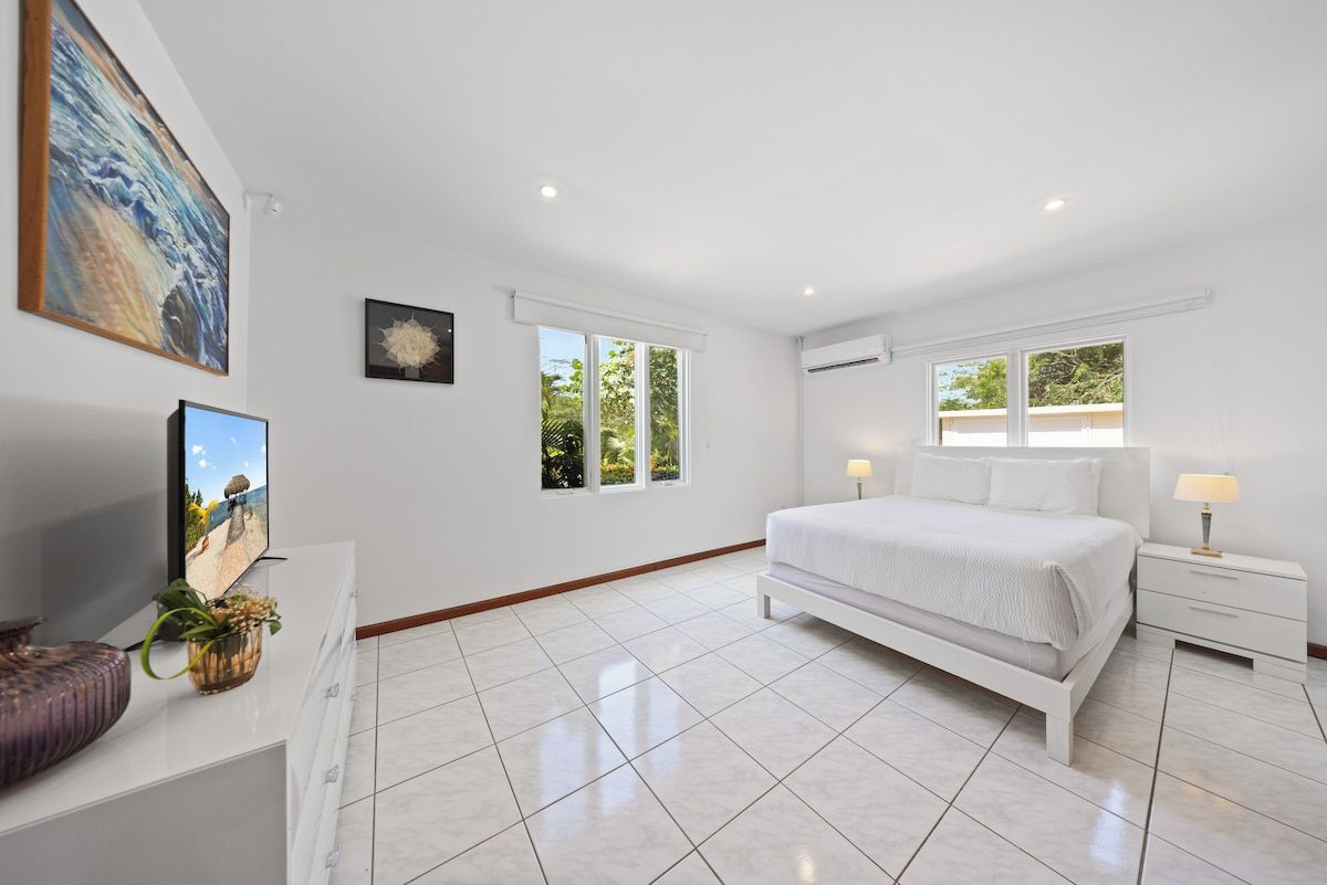 A modern bedroom with a large bed, a TV, and a window with a view of greenery.