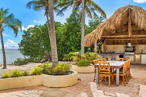 An outdoor kitchen with a thatched roof, surrounded by planters and a dining area with wooden chairs.