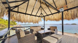 An outdoor seating area with wicker furniture and a thatched roof, overlooking a body of water.