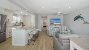 A modern kitchen with a central island, bar stools, and a dining area with a TV and striped armchairs.