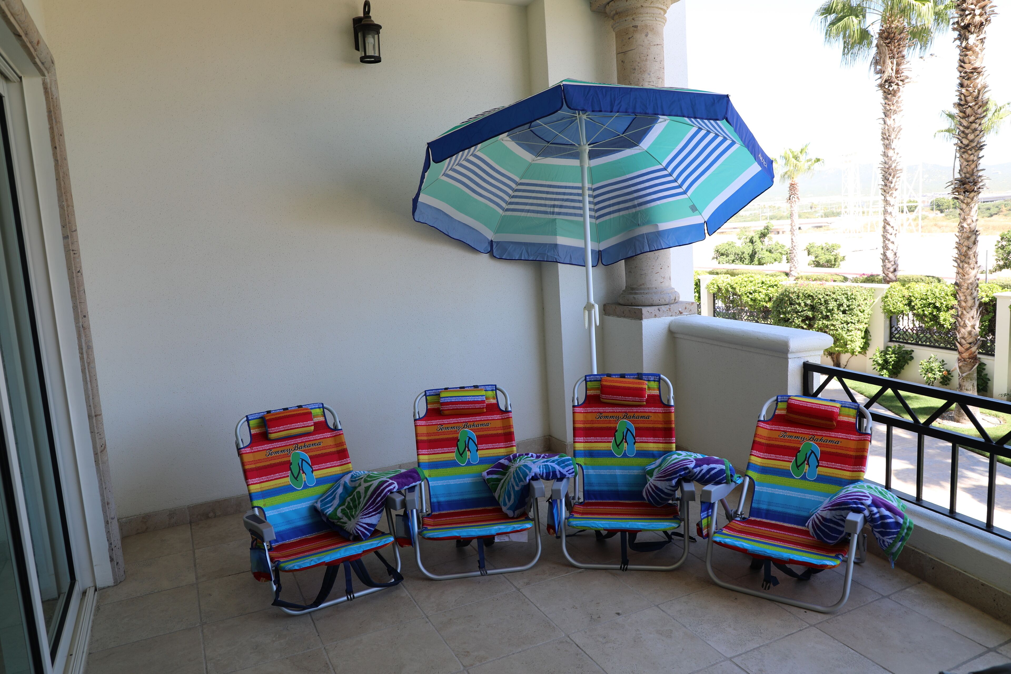 A balcony with four striped beach chairs, a blue and white striped umbrella, and a view of palm trees and a building in the background.