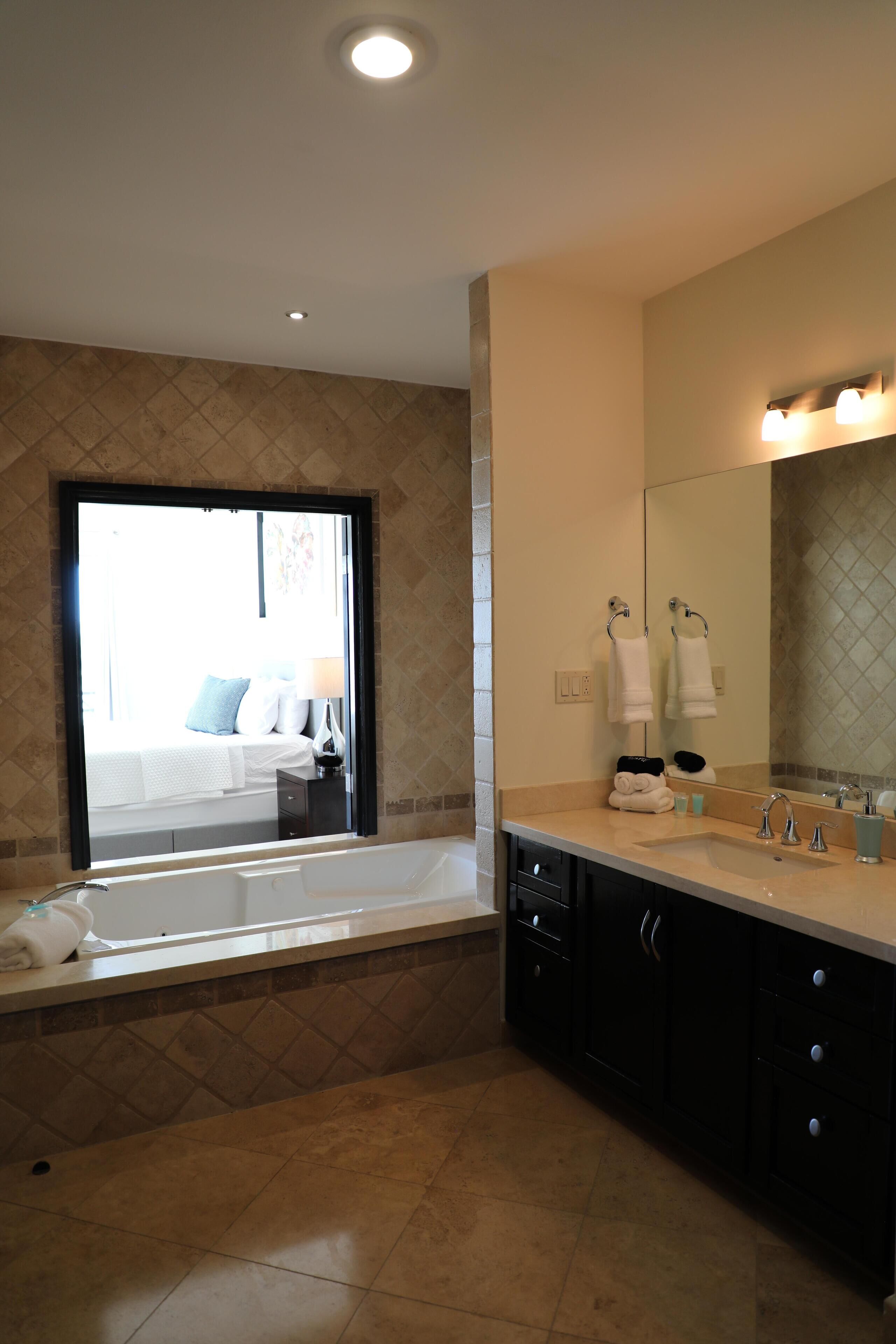 Une salle de bain moderne avec une baignoire, un grand miroir et une fenêtre donnant sur une chambre.
