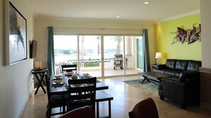 A spacious living area with a dining table, black leather sofa, and a view of palm trees through large glass doors.