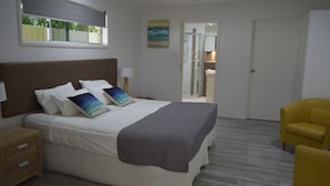 Ein Schlafzimmer mit Bett, Nachttischen, einem Sessel, einem Fenster mit Jalousien, einer Tür und einer Dusche.