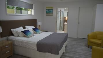 A bedroom with a bed, bedside tables, a chair, a window with blinds, a door, and a shower area.