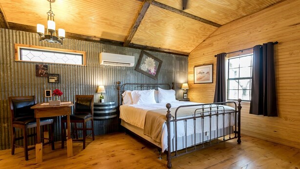 A cozy room with a wooden ceiling, a bed with white linens, a desk with a chair, and a window with curtains. - Bakery Haus- Red Velvet Suite | Wonderful Porch (Fredericksburg)
