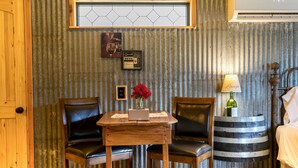 A rustic room with two leather chairs, a wooden table, a decorative wall with hexagonal tiles, a framed picture, and a lamp on a barrel. - Bakery Haus- Red Velvet Suite | Wonderful Porch (Fredericksburg)