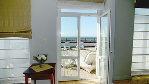 Una habitación con piso de madera, una mesa con un florero y una puerta corrediza de vidrio que da a un balcón con sillas blancas y vista al mar.