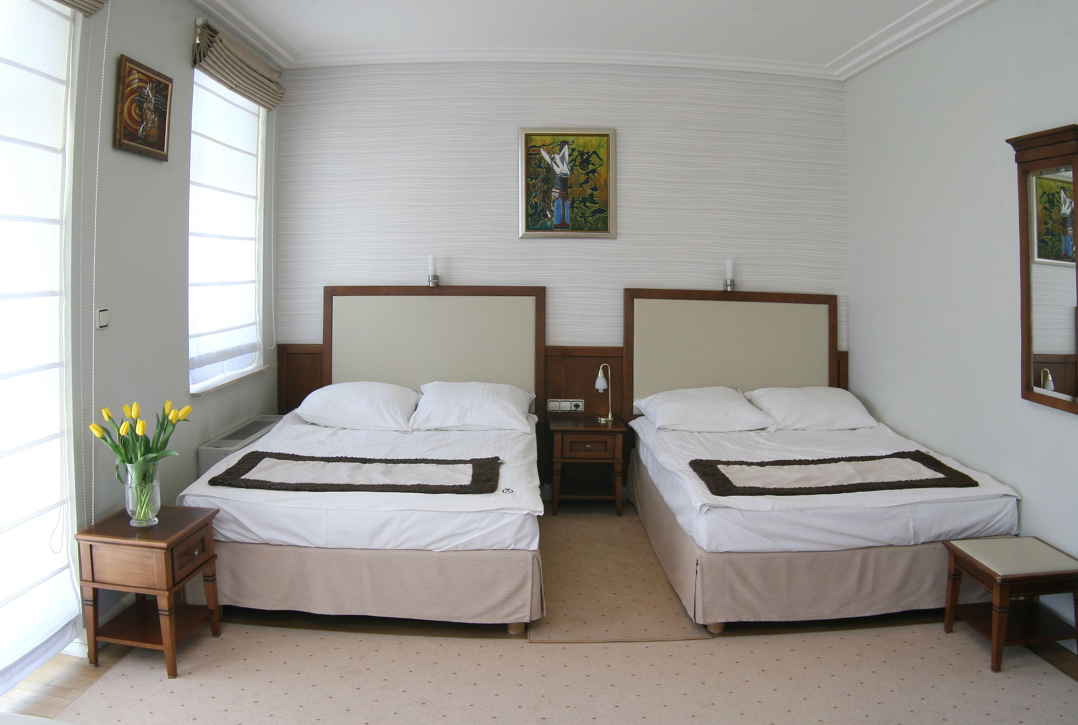 A double room with two beds, a nightstand, a bedside table, a vase with flowers, and a painting on the wall.
